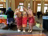 Three women wearing traditional Laotian dress hold up piece signs in front of a table and a background wall with tall rectangular windows.